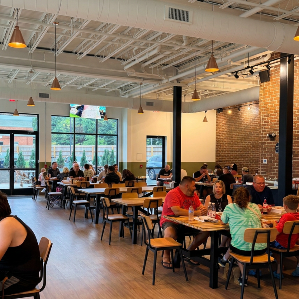 Busy casual restaurant dining room with exposed brick walls, pendant lighting, large windows, and patrons seated at wooden tables.