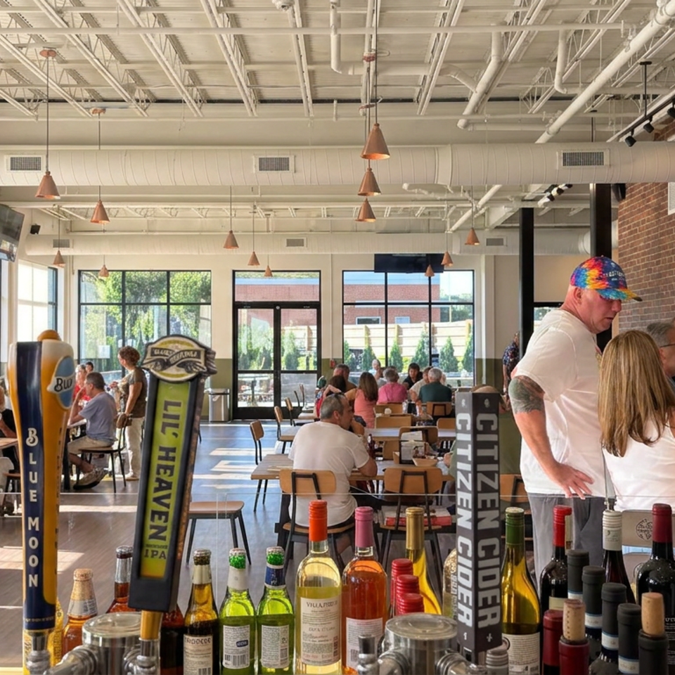 Bright restaurant interior with exposed brick, seated diners, standing patrons, beer taps, and assorted liquor bottles in the foreground.